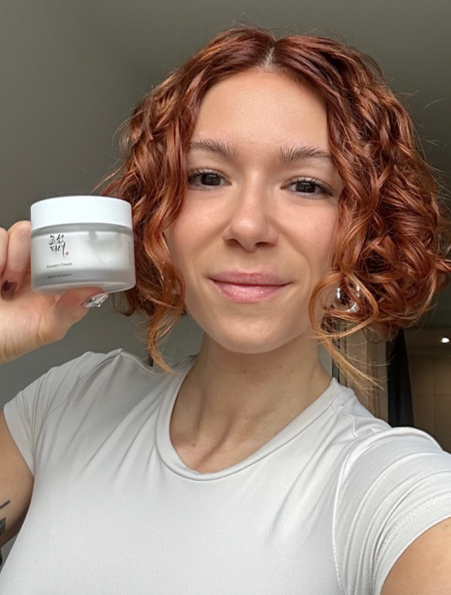 Someone holding a white pot of moisturiser with clear, dewy, healthy-looking skin