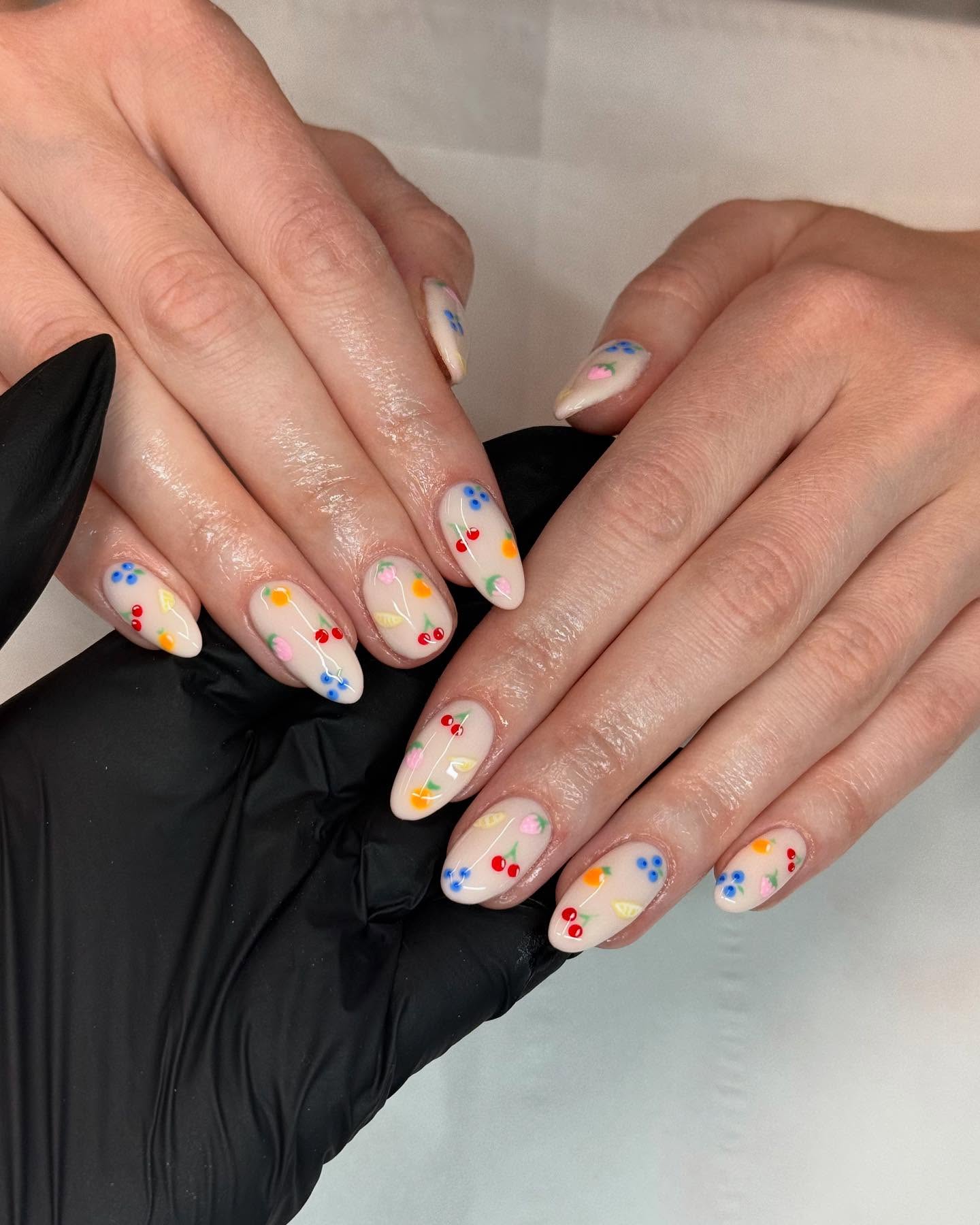 A close up of a hand with almond shaped nails featuring a neutral base colour and fruit designs