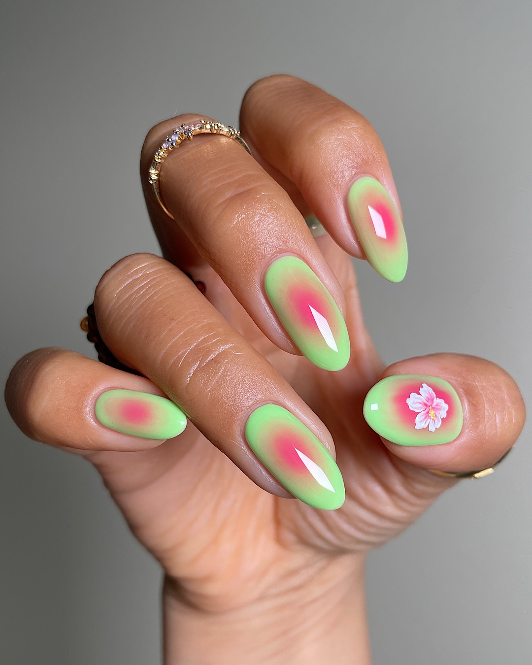 Close up of a hand with almond shaped nails featuring green nail varnish with a red gradient of colour in the middle and a white flower design on the thumb nail.