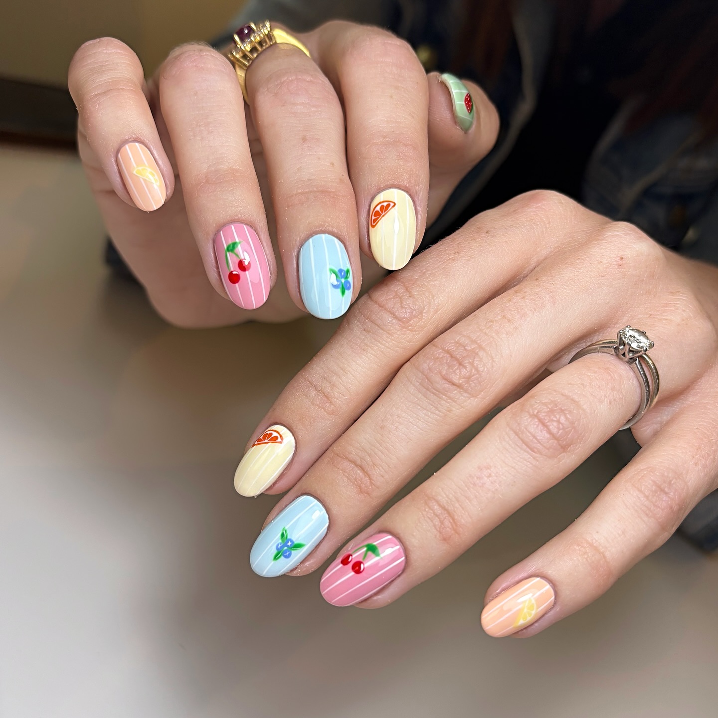A close up of a hand with almond shaped nails featuring yellow, blue, pink, and orange nails with small fruit designs